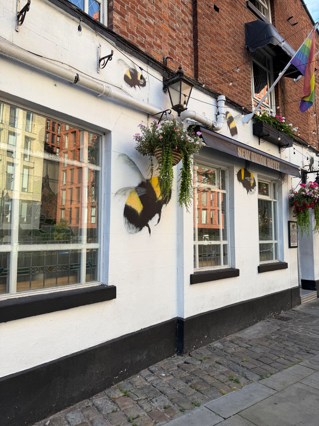 A whitewashed pub covered in large painted bumblebees, with hanging flower baskets and a rainbow flag