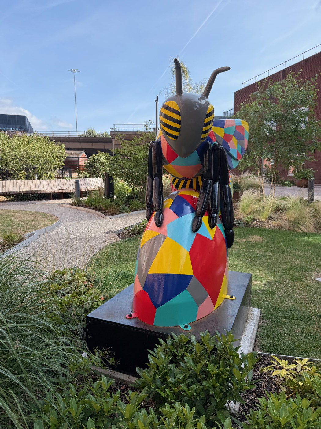 A large sculpture of a bee painted in bright geometric patchwork, standing in a landscaped city garden