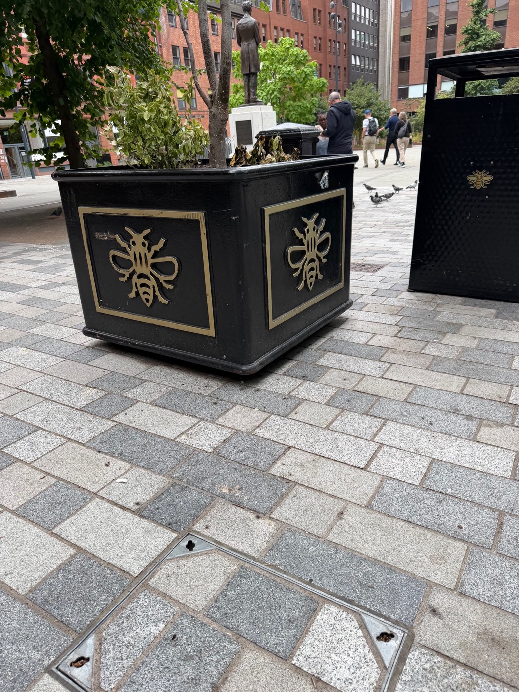A black planter and litter bin stamped with golden worker-bee emblems on a paved city square, pigeons in the background